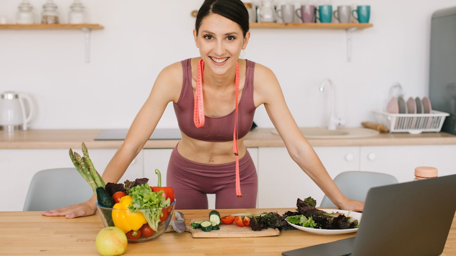 Smiling woman in athletic wear leans over a kitchen counter with a pink measuring tape draped around her neck, preparing a colorful vegetable salad in bowls.